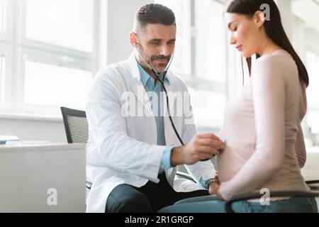 Donna incinta felice visita medico, medico maschile che esamina donna sorridente incinta, ascoltando il battito cardiaco del bambino Foto Stock