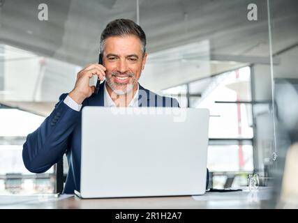 Felice uomo d'affari di mezza età impegnato che parla al telefono utilizzando un computer portatile in ufficio. Foto Stock