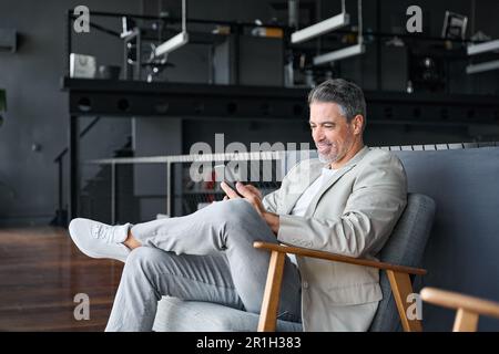 Uomo d'affari felice e maturo seduto sulla sedia in ufficio utilizzando il telefono cellulare. Foto Stock