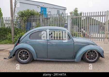 Rayleigh, Regno Unito. 14 maggio 2023. Un classico Maggiolino Volkswagen grigio-blu opaco con sospensione abbassata e ruote personalizzate parcheggiate accanto a un edificio industriale a Rayleigh, Essex. La giustapposizione di design vintage e modifiche moderne riflette la sottocultura automobilistica britannica in un ambiente urbano. Camion Farm Fresh Garage e veicoli americani in mostra. Penelope Barritt/Alamy Live News Foto Stock