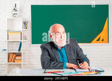 Ritratto di felice insegnante senior seduto in classe. Insegnante allegro che ama il suo lavoro. Divertente vecchio professore. Insegnante sorridente. Foto Stock