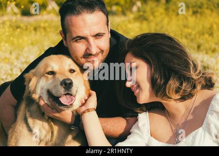 Su una coperta da picnic bianca, una donna incinta e suo marito condividono un tenero momento, mentre il loro cane amichevole cerca affetto nelle vicinanze Foto Stock