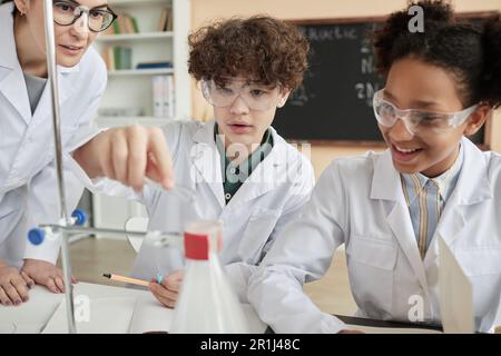 Ritratto di un ragazzo che fa esperimenti scientifici in classe scolastica con un gruppo di bambini che indossano camici da laboratorio Foto Stock