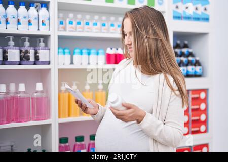 Giovane donna incinta cliente che legge la prescrizione tenendo pillole bottiglia in farmacia Foto Stock