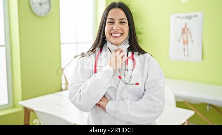 Giovane donna ispanica bella medico che prende la maschera medica sorridendo in clinica Foto Stock