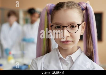 Primo piano ritratto di ragazza scolastica carina teen con pigtail vestito da laboratorio in classe scientifica e guardando la fotocamera con espressione seria del volto Foto Stock