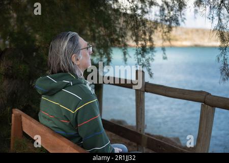 Un uomo con lunghi capelli grigi e bicchieri seduti su una panchina solo sul mare, Grecia, Creta Foto Stock