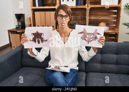 Donna brunette che lavora con rorschach test a psicologia clinica sticking lingua fuori felice con l'espressione divertente. Foto Stock