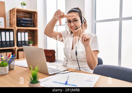 Giovane donna ispanica che indossa la cuffia del call center in ufficio sorridendo rendendo la cornice con le mani e le dita con il viso felice. creatività e foto Foto Stock