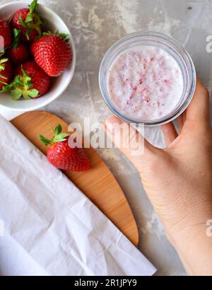 Vista dall'alto della tazza di vetro con frullato di fragole. La mano di una donna tiene un bicchiere di frullato. Su sfondo grigio, un piattino con fragole fresche. Foto Stock
