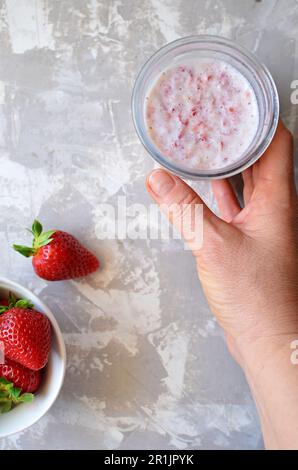 Vista dall'alto della tazza di vetro con frullato di fragole. La mano di una donna tiene un bicchiere di frullato. Su sfondo grigio, un piattino con fragole fresche. Foto Stock