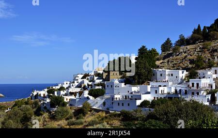 Bianche case medievali sulle pendici di una collina nella baia del villaggio di Lindos a Rodi. Mare blu ai piedi della collina e gli alberi tra le case. Foto Stock