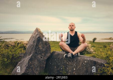 Splendido scenario naturalistico con uomo in camicia senza maniche e pantaloncini camo meditati sulla roccia sul lago Corrib a Galway, Irlanda Foto Stock