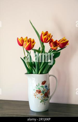 Caraffa in porcellana bianca con stampa floreale contenente un bouquet di tulipani rossi e gialli su una mensola di legno. Immagine verticale con messa a fuoco e copia selettive Foto Stock