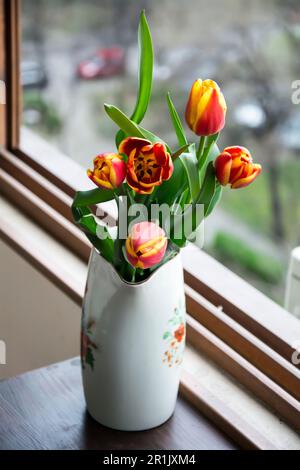 Una caraffa di porcellana bianca contenente un bouquet di tulipani rossi e gialli accanto alla finestra. Immagine verticale con messa a fuoco selettiva e sfondo sfocato. Foto Stock