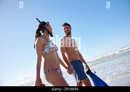 Sole, mare e relax. Inquadratura ad angolo basso di una giovane coppia che cammina lungo una spiaggia insieme all'attrezzatura da snorkeling. Foto Stock