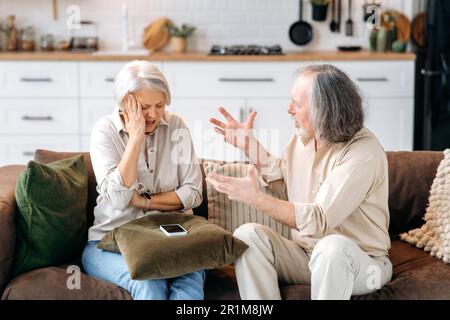 Coppia caucasica matura, pensionati, sedersi sul divano in soggiorno, litigare tra loro, gridare, giurare, provare irritazione e indignazione, dissentire tra loro, gesticulare con le mani Foto Stock