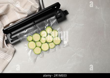 Sigillante per confezionamento sottovuoto e sacchetto di plastica con zucchine tagliate su tavolo in marmo grigio chiaro, vista dall'alto. Spazio per il testo Foto Stock