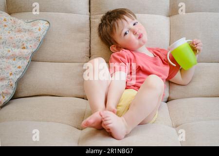 La tazza antigoccia è nelle mani del bambino sorridente, che siede contentedly sul divano, bevendo acqua. Un bambino beve liquido da una p verde Foto Stock