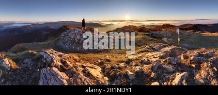 Alba con inversione sopra la profonda valle autunnale delle montagne con piccolo uomo in cima, che sta guardando questo bellissimo paesaggio Foto Stock