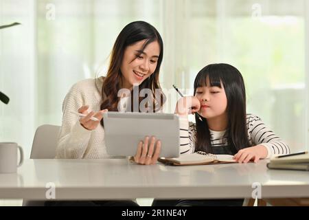 Piccola ragazza asiatica che studia, guarda una lezione virtuale online su un tablet digitale con la madre durante la formazione remota Foto Stock