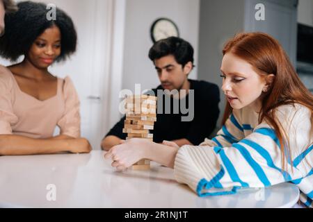 Vista laterale di una giovane donna rossa focalizzata che prende con attenzione il blocco di legno jenga dal fondo della torre mentre gioca con gli amici durante la festa Foto Stock