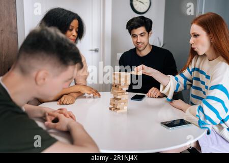 Giovane donna dai capelli rossi focalizzata che rimuove con attenzione il blocco di legno dalla struttura di torre di legno. Concentrata bella donna seduta a tavola che gioca la società Foto Stock