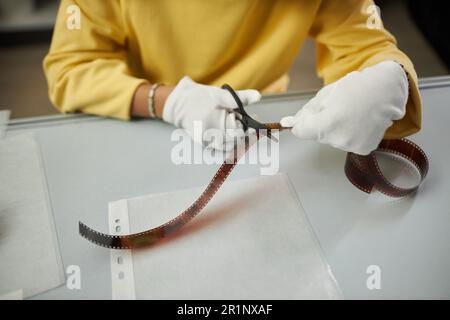 Primo piano del fotografo in guanti bianchi che taglia la pellicola fotografica con le forbici mentre si siede al suo posto di lavoro Foto Stock