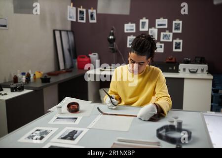 Giovane fotografo seduto sul suo posto di lavoro e tagliare un film fotografico in studio Foto Stock