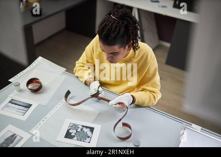 Vista ad alto angolo del giovane fotografo che taglia un film fotografico seduto sul suo posto di lavoro in studio fotografico Foto Stock