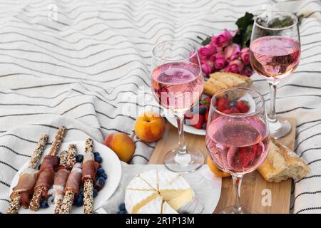 Bicchieri di delizioso vino rosa, fiori e cibo su coperta bianca da picnic Foto Stock