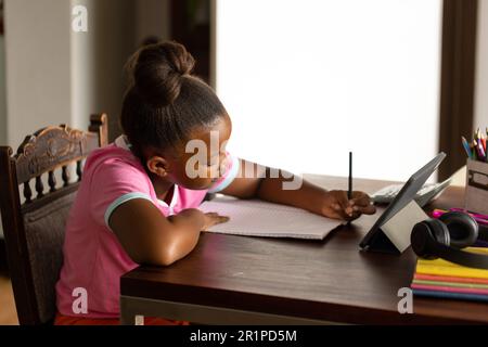 Felice ragazza afroamericana seduta al tavolo da pranzo facendo scuola utilizzando un tablet a casa Foto Stock