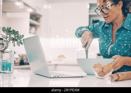 Felice donna adulta cottura torta panetteria a casa guardando tutorial istruzioni sul computer portatile sul tavolo. Moderno on-line cuoco femminile persone. Donna moglie persone che preparano il cibo al coperto in appartamento Foto Stock