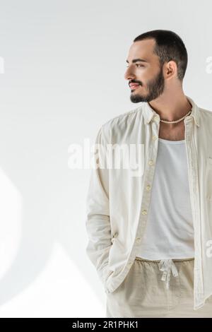 Uomo omosessuale bearded e short haired in camicia beige ed elegante collana in perla guardando lontano e tenendo la mano in tasca dei pantaloni fatti di fava naturale Foto Stock