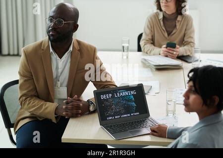 Giovane uomo d'affari serio ed i suoi colleghi che ascoltano la relazione dell'oratore mentre sono seduti alla scrivania durante una conferenza sull'apprendimento profondo Foto Stock