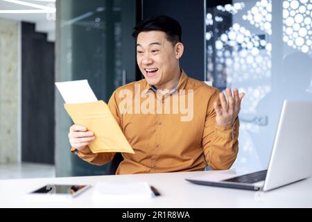 Felice uomo asiatico ha ricevuto la lettera apre e legge il documento del messaggio, l'uomo d'affari lavora all'interno di un moderno edificio di uffici sul laptop, buone notizie nella notifica. Foto Stock