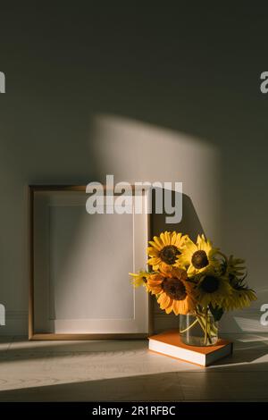 Vaso di girasoli su un libro hardback accanto a una cornice vuota nella luce del sole Foto Stock