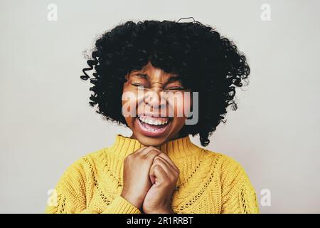 Primo piano ritratto di allegra donna nera ridendo con gli occhi chiusi tenendo le mani indossando maglione giallo isolato su sfondo grigio Foto Stock