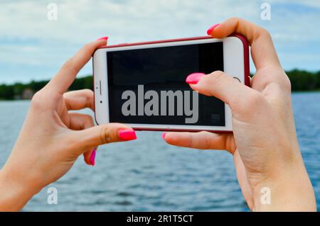Una donna ragazza con belle unghie manicure rosa arancione tiene in mano uno smartphone cellulare e scatta fotografie della natura acqua il fiume se Foto Stock