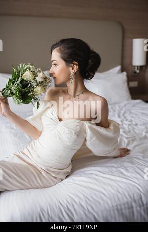 giovane sposa in abito bianco e gioielli di lusso seduti sul letto e odore bouquet nuziale con fiori mentre si guarda lontano nella moderna camera da letto dell'hotel Foto Stock