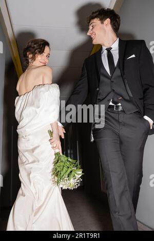 vista dal basso angolo dello sposo allegro in abito nero posa con mano in tasca e guardando la sposa felice in abito da sposa bianco con bouquet nuziale whi Foto Stock