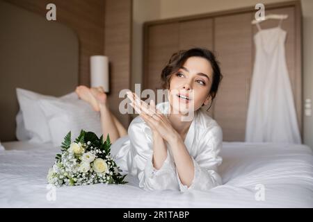 splendida donna con capelli brunette e anello di fidanzamento sul dito guardando lontano e sdraiato in bianco vestito di seta accanto al bouquet nuziale sul letto con matrimonio d Foto Stock