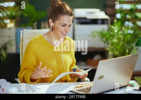 sorridente elegante piccola impresa proprietaria donna in maglione giallo con portatile che ha riunione virtuale nel moderno ufficio verde. Foto Stock