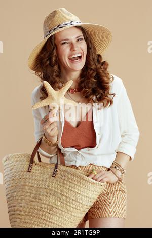 Vacanza in spiaggia. felice casalinga elegante in blusa bianca e pantaloncini su sfondo beige con stella marina, borsa in paglia e cappello estivo. Foto Stock