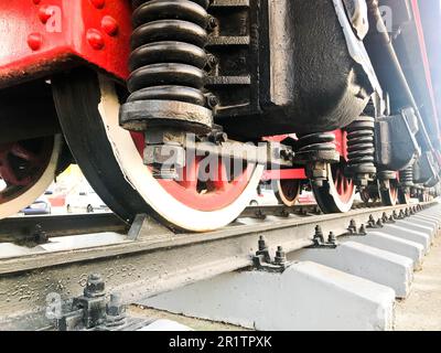 Grandi ruote di ferro di un treno rosso e nero su rotaie ed elementi di sospensione con molle di una vecchia locomotiva industriale a vapore. Foto Stock