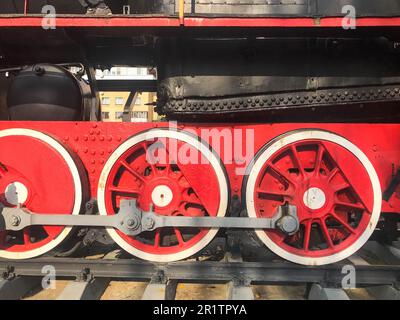 Grandi ruote di ferro di un treno rosso e nero su rotaie ed elementi di sospensione con molle di una vecchia locomotiva industriale a vapore. Foto Stock