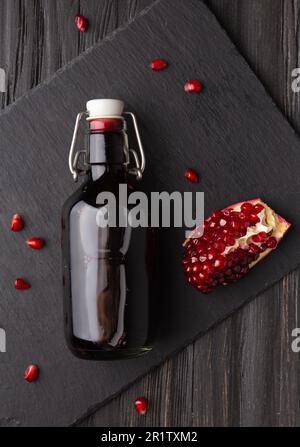 Spremuta di melograno in una bottiglia trasparente di vetro, melograno spezzato e semi di melograno su un piatto di pietra piatta nera Foto Stock
