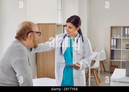 Giovane, bella, sorridente donna medico in cappotto bianco rispettosamente comunica con l'uomo anziano in occhiali dando voce alle notizie positive nell'ufficio di Foto Stock