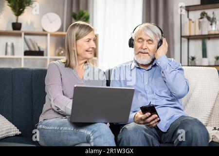 Allegra coppia senior che si diverte su un comodo divano utilizzando un moderno computer portatile per guardare lo spettacolo comico in un elegante salotto. Donna sorridente che regola gli auricolari wireless per il marito amato. Foto Stock