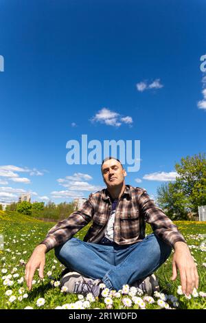 Un giovane bell'uomo è seduto e si diverte in un prato di fiori soleggiato in una giornata di sole. Giovane uomo rilassato seduto in un campo di fiori Foto Stock